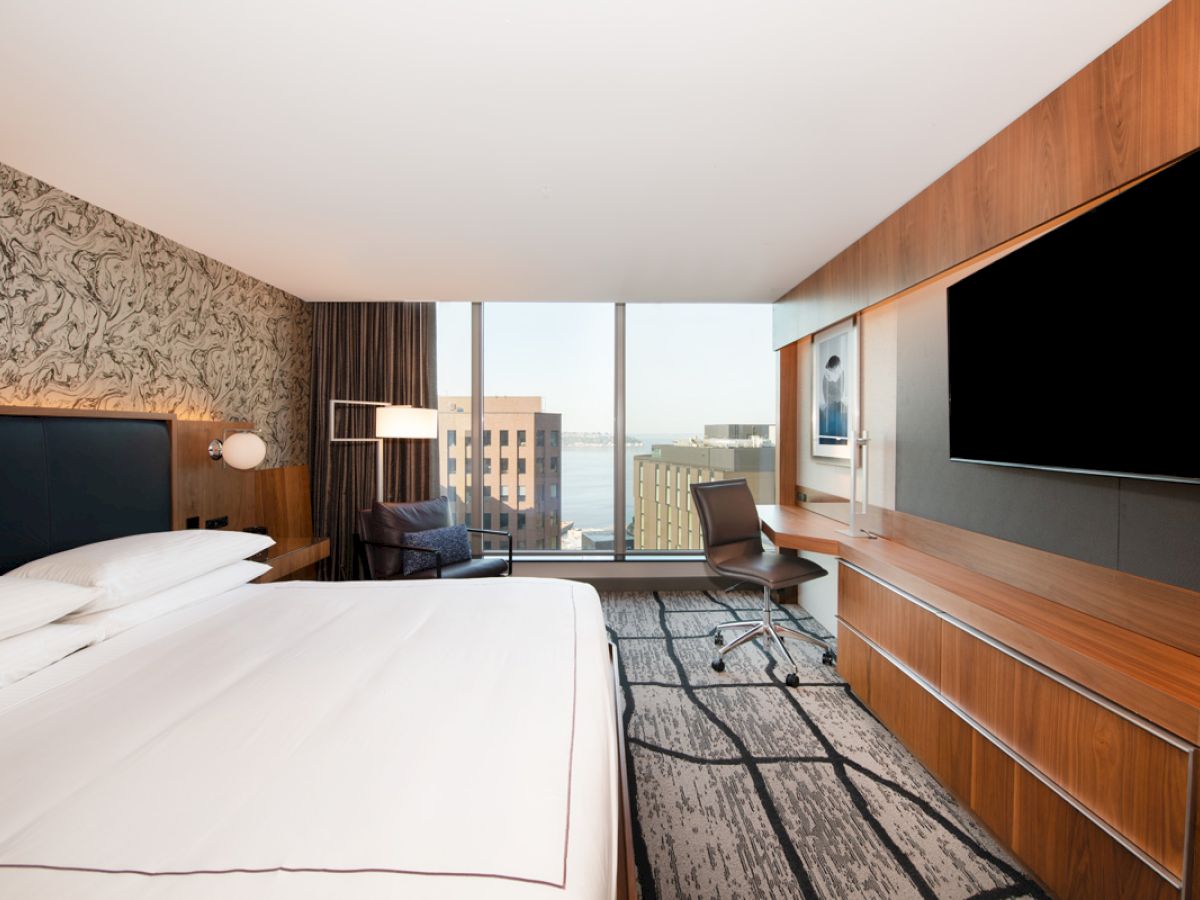 A modern hotel room with a large bed, wall-mounted TV, desk, and chair, featuring a city view through a floor-to-ceiling window.