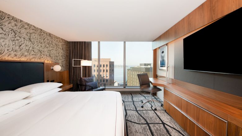 A modern hotel room with a large bed, wall-mounted TV, desk, and chair, featuring a city view through a floor-to-ceiling window.
