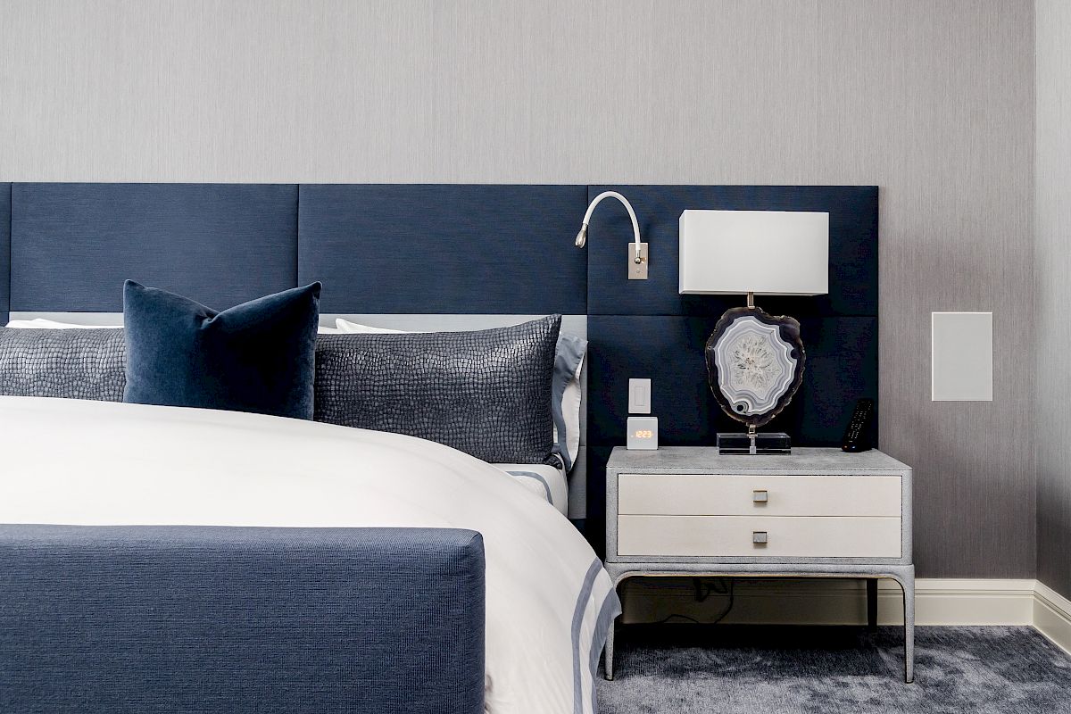 A modern bedroom with a blue headboard, side table with a lamp and decor, grey walls, and carpeting.