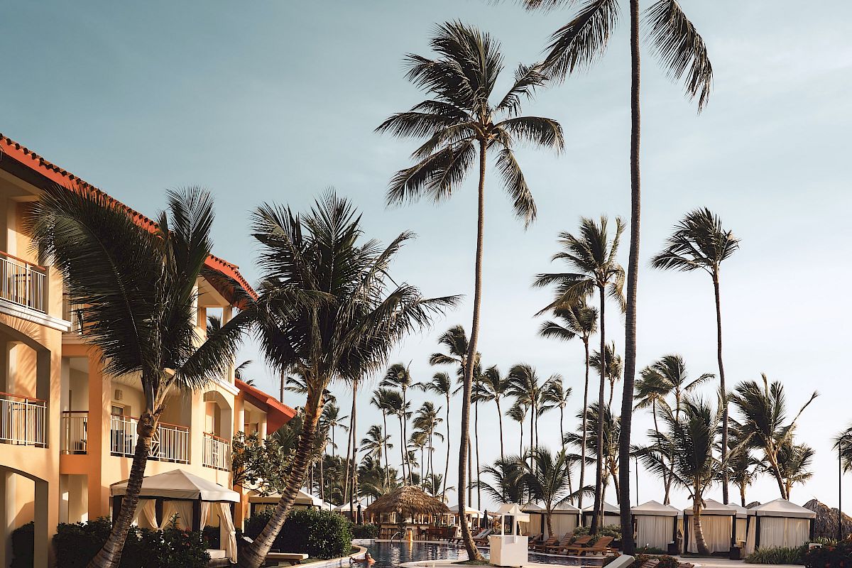 A serene luxury resort poolside area with lounge chairs, tall palm trees, a clear blue sky, and part of a building with balconies in the background.