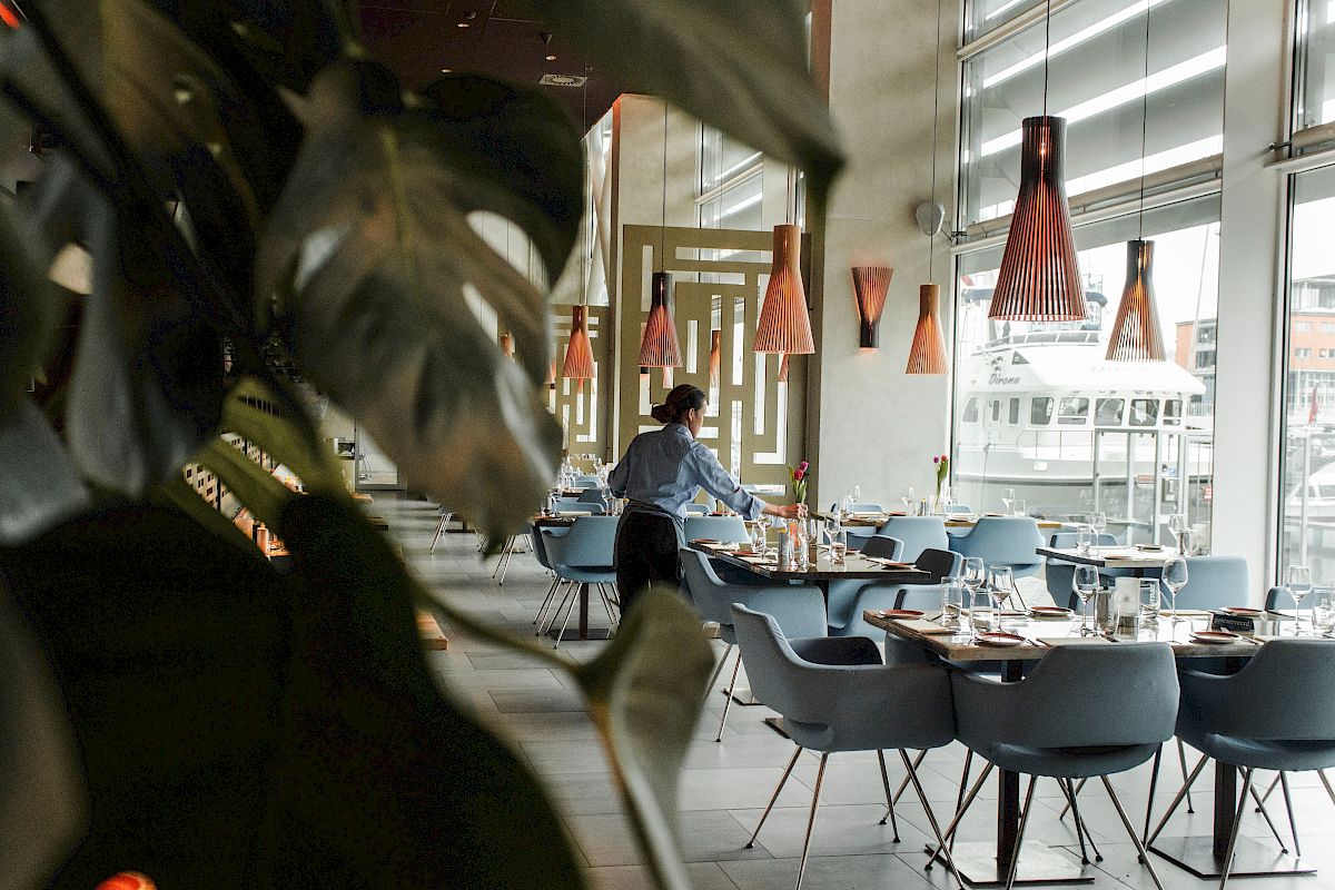 The image depicts a modern, well-lit restaurant where a person is setting up tables, with large windows and stylish pendant lights overhead.