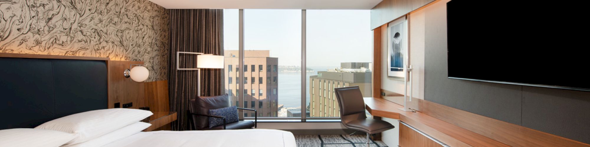The image shows a modern hotel room with a king-size bed, a large TV, a desk and chair, and a view of buildings and water through the window.