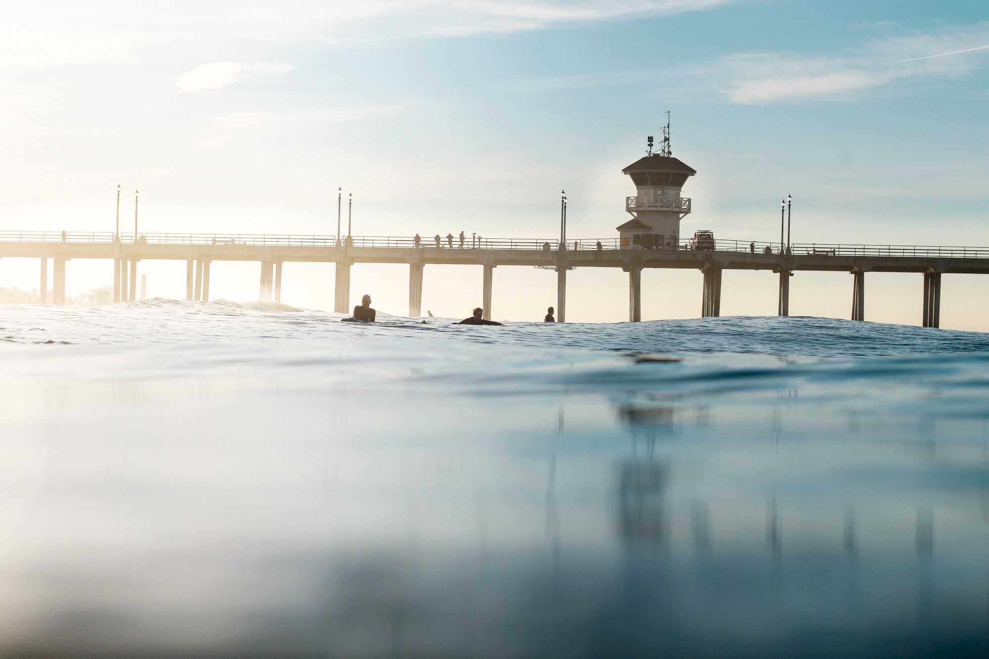 Surfers and a pier are visible above calm ocean waters on a sunny day, with a tower-like structure on the pier.